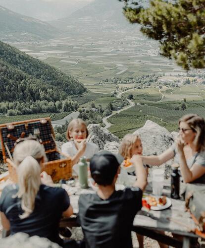 Several adults and children are sitting at a wooden table outdoors, with a filled picnic basket on it; the Vinschgau valley landscape is visible in the sunshine in the background.