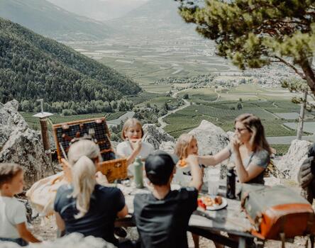 Several adults and children are sitting at a wooden table outdoors, with a filled picnic basket on it; the Vinschgau valley landscape is visible in the sunshine in the background.
