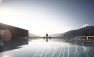 Piscina a sfioro con vista sulle montagne all’alba o al tramonto; una persona è seduta sul bordo della piscina, la superficie calma dell’acqua riflette il cielo.