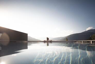 Infinity pool with a view of the mountains at sunrise or sunset; a person is sitting on the pool edge, calm water surface reflecting the sky.