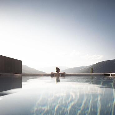 Infinity pool with a view of the mountains at sunrise or sunset; a person is sitting on the pool edge, calm water surface reflecting the sky.