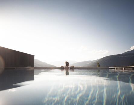 Infinity-Pool mit Blick auf die Berge bei Sonnenaufgang oder Sonnenuntergang; eine Person sitzt am Beckenrand, ruhige Wasseroberfläche reflektiert den Himmel.