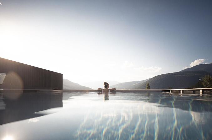 Infinity-Pool mit Blick auf die Berge bei Sonnenaufgang oder Sonnenuntergang; eine Person sitzt am Beckenrand, ruhige Wasseroberfläche reflektiert den Himmel.