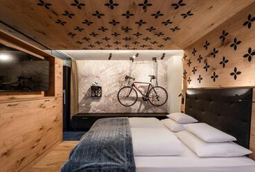Camera d’hotel moderna con letto matrimoniale, pareti in legno e televisore; sulla parete di fondo sono appesi una bicicletta, scarpe da ciclismo e un casco da bici.