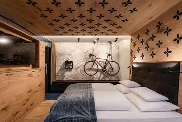 Modernes Hotelzimmer mit Doppelbett, Holzwänden und Fernseher; an der Rückwand hängen ein Fahrrad, Fahrradschuhe und ein Fahrradhelm.