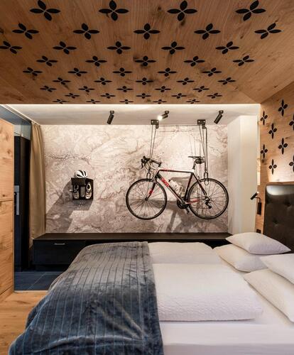 Camera d’hotel moderna con letto matrimoniale, pareti in legno e televisore; sulla parete di fondo sono appesi una bicicletta, scarpe da ciclismo e un casco da bici.