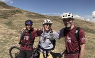 Three bikers wearing helmets stand amidst expansive alpine meadows; blue sky in the background.