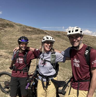 Tre biker con il casco si trovano in mezzo a vasti prati alpini; sullo sfondo cielo azzurro