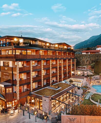 An exterior shot of the hotel, including the pool area with loungers on the sun lawn in an evening setting; hilly landscape and blue sky in the background.