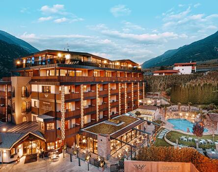 An exterior shot of the hotel, including the pool area with loungers on the sun lawn in an evening setting; hilly landscape and blue sky in the background.