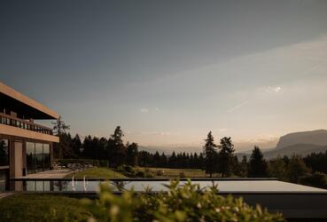Im Vordergrund ein Pool mit Liegewiese im Sonnenuntergang daneben ist ein Teil des modernen Hotel Pfösl zu sehen; im Hintergrund Wald, Hügel und Berglandschaft.