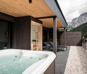 Whirlpool auf einer Hotelterrasse mit Glasfront und Holzboden; im Hintergrund sind Berge und bewaldete Hänge zu sehen.