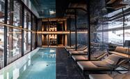 Interno di un'area benessere moderna con piscina interna, lettini sul lato destro, grandi finestre in vetro e pavimento scuro.