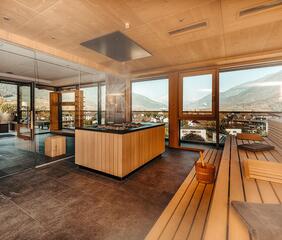 Interior view of the sauna area with wooden benches, large panoramic windows, a view of the mountain landscape and natural daylight; a sauna stove with chimney in the center.