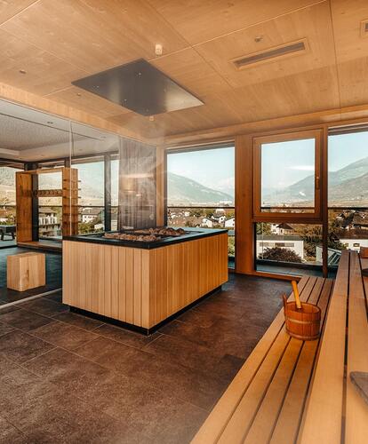 Interior view of the sauna area with wooden benches, large panoramic windows, a view of the mountain landscape and natural daylight; a sauna stove with chimney in the center.
