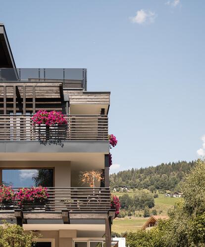 Vista esterna del Bikehotel Mesnerwirt ad Avelengo; balconi in legno con fiori, un’ampia facciata vetrata; sullo sfondo bosco, prati, montagne e cielo estivo azzurro.
