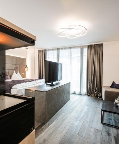 Modern hotel room with seating area, sofa, coffee table, panoramic windows, and built-in fireplace; bright daylight streaming through the windows.