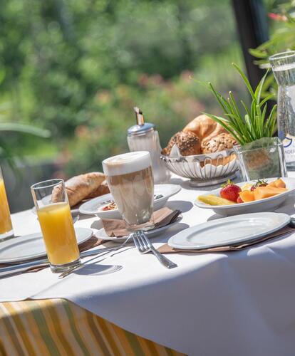 Tavolo da colazione apparecchiato per due persone; sul tavolo ci sono due cappuccini, un piatto di frutta, due bicchieri di succo, una brocca d’acqua, un cestino di pane, croissant, uno zuccheriere e una pianta verde.