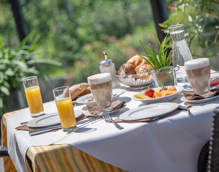 Tavolo da colazione apparecchiato per due persone; sul tavolo ci sono due cappuccini, un piatto di frutta, due bicchieri di succo, una brocca d’acqua, un cestino di pane, croissant, uno zuccheriere e una pianta verde.