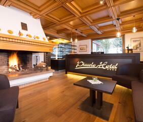 Lobby of the Piccolo Hotel with reception, wooden ceiling and lit fireplace; in the foreground, there are armchairs and a small table.