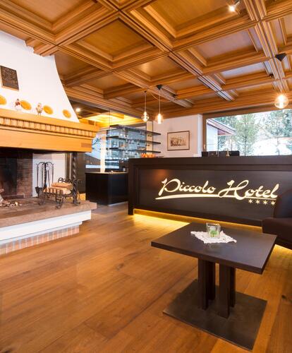 Lobby of the Piccolo Hotel with reception, wooden ceiling and lit fireplace; in the foreground, there are armchairs and a small table.