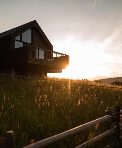 Modernes Holzhaus auf einer grünen Wiese bei Sonnenuntergang, mit Holzzaun im Vordergrund und bewaldeten Hügeln im Hintergrund.