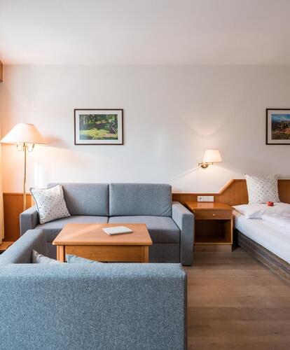 Camera d'hotel luminosa con due letti singoli, divano grigio, comodini con lampade, grande finestra con tende e tavolo con sedie.