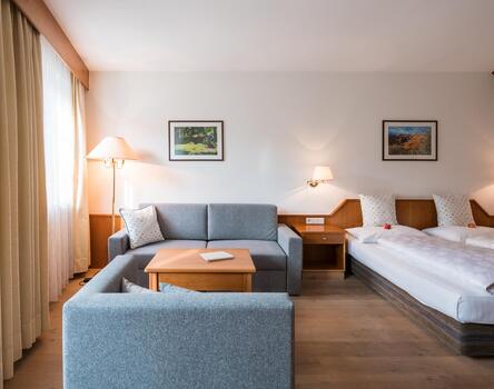 Camera d'hotel luminosa con due letti singoli, divano grigio, comodini con lampade, grande finestra con tende e tavolo con sedie.