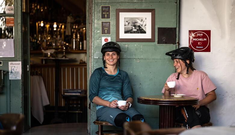 Two women wearing bicycle helmets drink a coffee