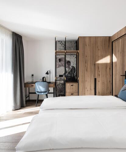 Camera d'hotel moderna con letto matrimoniale, cuscini grigi, grande finestra con tende, TV a schermo piatto e area lavoro sullo sfondo.