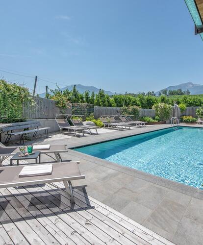 Foto diurno di un hotel con area piscina, terrazza e lettini; sullo sfondo alberi da frutto e cielo sereno.