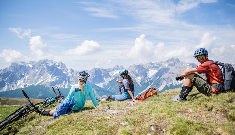 A man and two women are sitting on an alpine meadow and looking at the Dolomites. Their bicycles are lying next to them