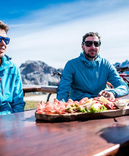 Two bikers in blue jackets are sitting at a wooden table outdoors; on the table there is a platter of food, with bicycles, helmets and mountains visible in the background.