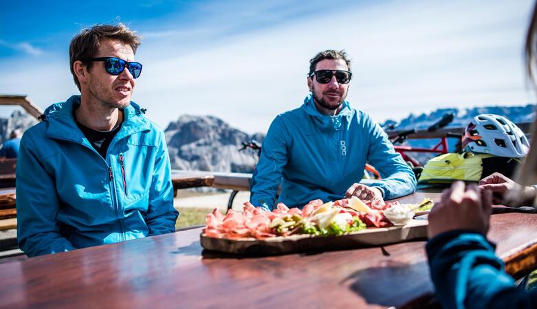 Two bikers in blue jackets are sitting at a wooden table outdoors; on the table there is a platter of food, with bicycles, helmets and mountains visible in the background.