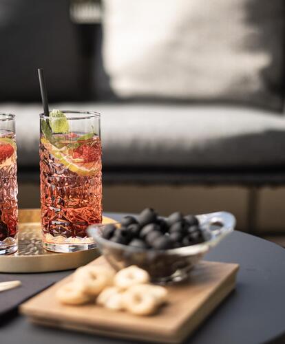 Primo piano di un piccolo tavolo rotondo con due aperitivi e snack; sullo sfondo una poltrona in vimini imbottita.