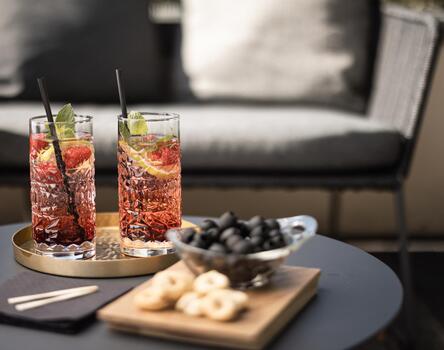 Primo piano di un piccolo tavolo rotondo con due aperitivi e snack; sullo sfondo una poltrona in vimini imbottita.