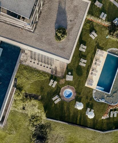 Luftaufnahme des Hotel-Außenbereichs mit Pool, Liegewiese, Sonnenschirmen und angrenzender Berglandschaft.