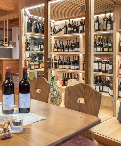Scaffale di vini all’interno con tavoli e sedie in legno; su un tavolo ci sono due bottiglie di vino e bicchieri.