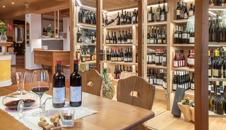 Scaffale di vini all’interno con tavoli e sedie in legno; su un tavolo ci sono due bottiglie di vino e bicchieri.