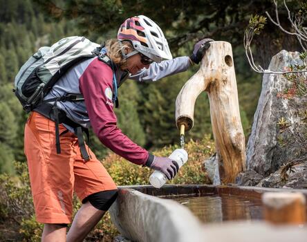 Ciclista con pantaloni rossi e casco che attinge acqua da un pozzo di legno nella natura