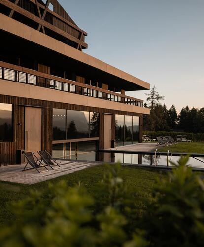 Das moderne Hotel Pfösl mit Außenpool und Liegewiese im Sonnenuntergang; im Hintergrund Wald und Berge.