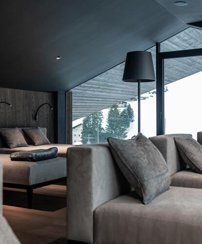 Primo piano di un'area benessere arredata in stile moderno con lettini grigio scuro, cuscini e lampade da terra scure davanti a una vetrata.