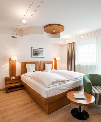 Camera d'hotel moderna e luminosa con letto matrimoniale, lampade da comodino, una poltrona, un piccolo tavolo rotondo e una grande vetrata.