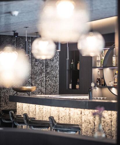 Modern hotel bar area with illuminated counter, high stools, and multiple hanging lamps; blurred foreground for atmospheric effect.