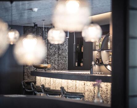 Moderner Hotelbarbereich mit beleuchtetem Tresen, hohen Hockern und mehreren hängenden Lampen, unscharfer Vordergrund für atmosphärische Wirkung.