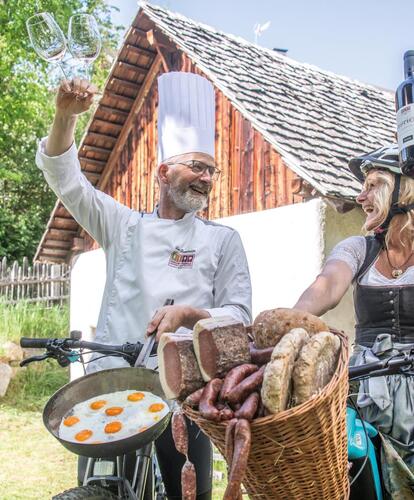 Petra e Andreas brindano sulle loro biciclette con un bicchiere di vino rosso. Tengono un cesto con pane, salumi e pancetta