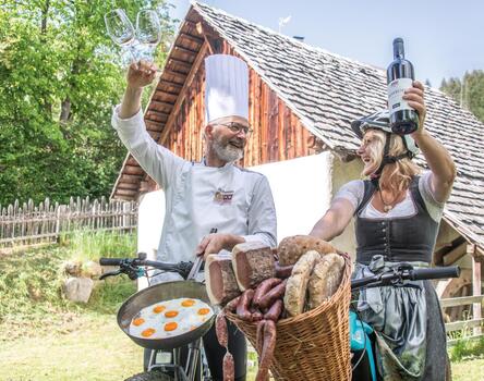 Petra e Andreas brindano sulle loro biciclette con un bicchiere di vino rosso. Tengono un cesto con pane, salumi e pancetta