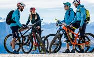 Vier Biker mit ihren Fahrrädern und Helmen stehen auf einem Weg zusammen; im Hintergrund ist die massive Berglandschaft zu sehen.