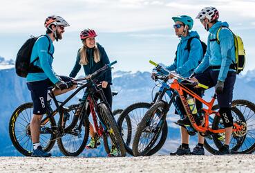 Four bikers with their bicycles and helmets stand together on a path; in the background, a massive mountain landscape can be seen.