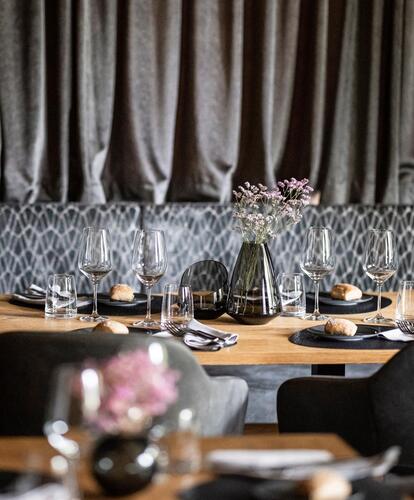 Sala da pranzo moderna con sedute rivestite in grigio scuro, tavoli in legno e una lunga tenda scura sullo sfondo; un tavolo apparecchiato per quattro persone con un vaso di fiori al centro.